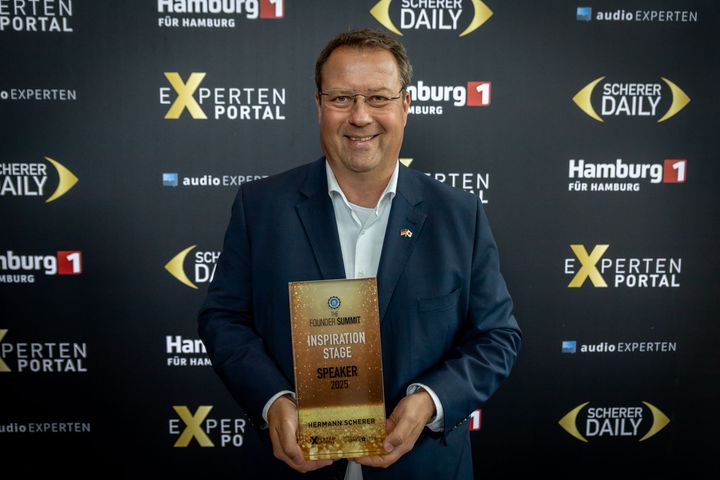 Ein Mann im Anzug hält eine Trophäe vor einer schwarzen Wand.