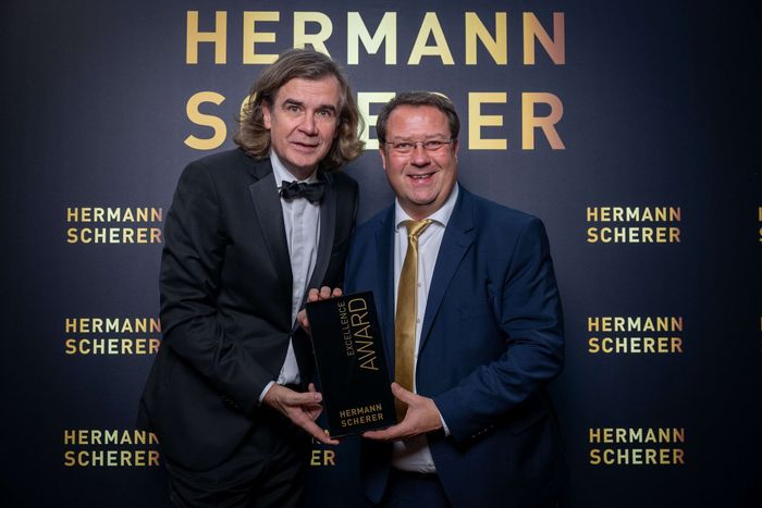 Zwei Männer in Anzügen stehen nebeneinander und halten eine Trophäe in den Händen.