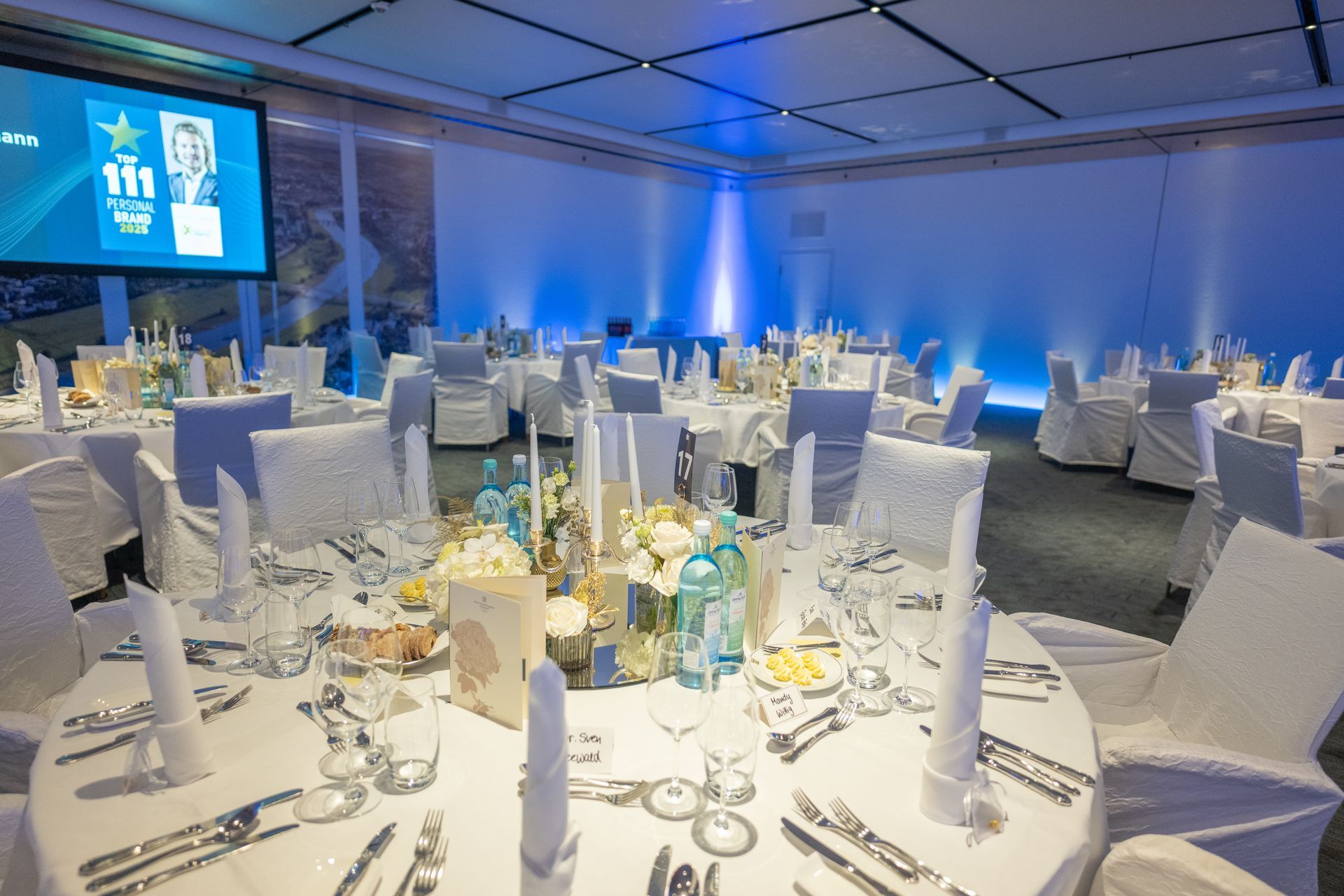 Ein formelles Esszimmer mit weiß gedeckten Tischen für ein Bankett. Die Tische sind mit Blumengestecken geschmückt, die mit blauem Licht beleuchtet sind.