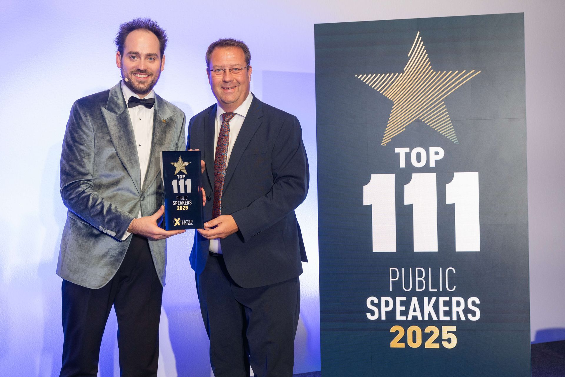 Birger Nispel und Josua Laufer halten auf der Bühne eine Auszeichnung in die Höhe, daneben ein Schild mit der Aufschrift „Top 111 Public Speakers 2025“.
