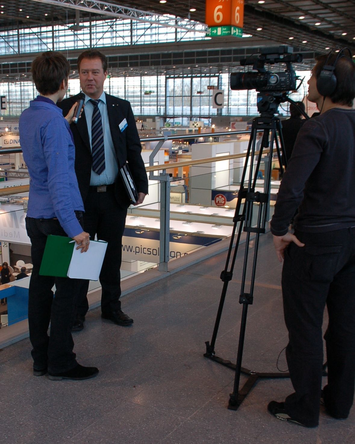 Ein Reporter interviewt einen Mann im Anzug, während ein Kameramann aufzeichnet. Drinnen, bei einer Veranstaltung.