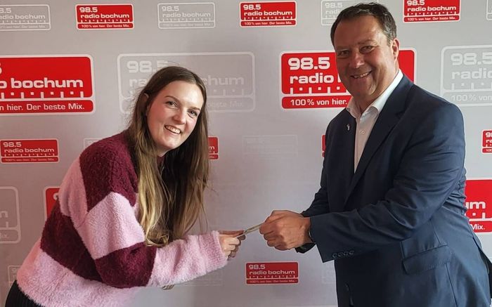 Ein Mann und eine Frau geben sich vor einer Wand mit der Aufschrift „98.5 Radio“ die Hand.