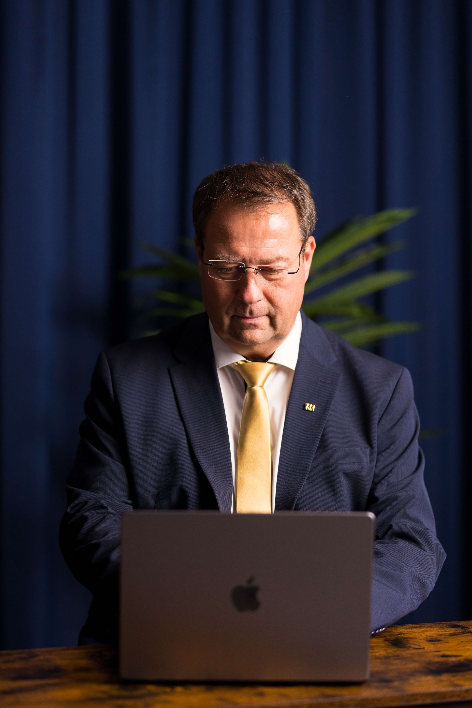 Birger Nispel im Anzug, mit Brille und gelber Krawatte benutzt einen Laptop vor einem blauen Vorhang.