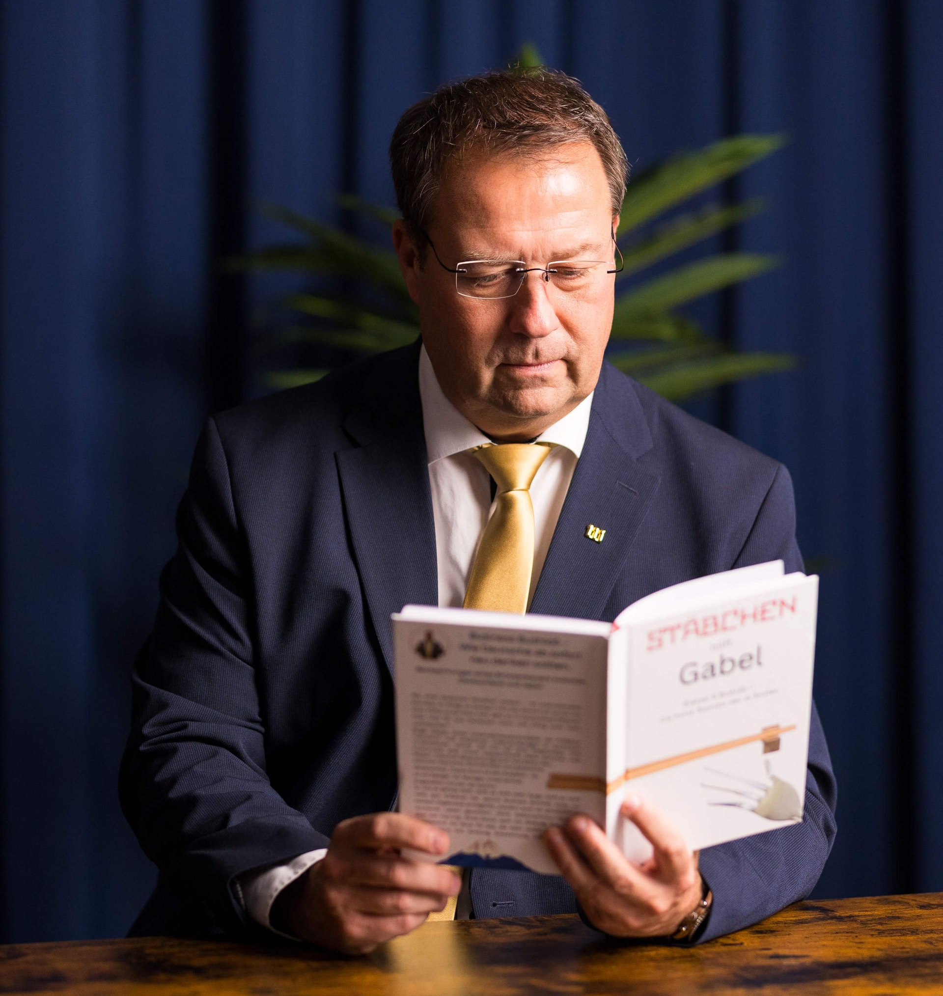 Birger Nispel im Anzug liest an einem Holztisch vor dunkelblauem Hintergrund das Buch „Gabel hilft Stäbchen“.