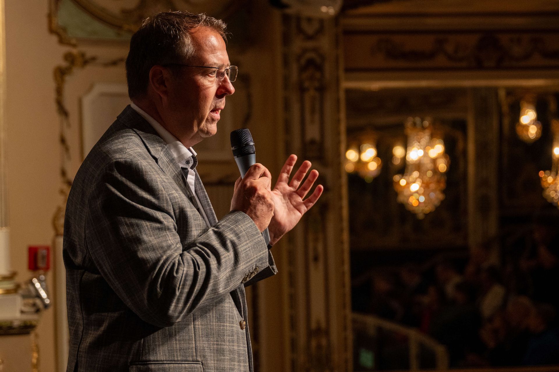 Ein Mann spricht in ein Mikrofon und gestikuliert mit den Händen. Er trägt einen Anzug und steht in einem eleganten Raum mit Kronleuchtern.