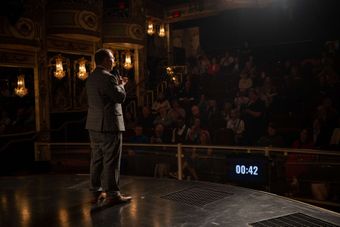 Mann auf der Bühne spricht zum Publikum in einem Theater. Dunkel, gut gekleidet; digitaler Timer sichtbar.