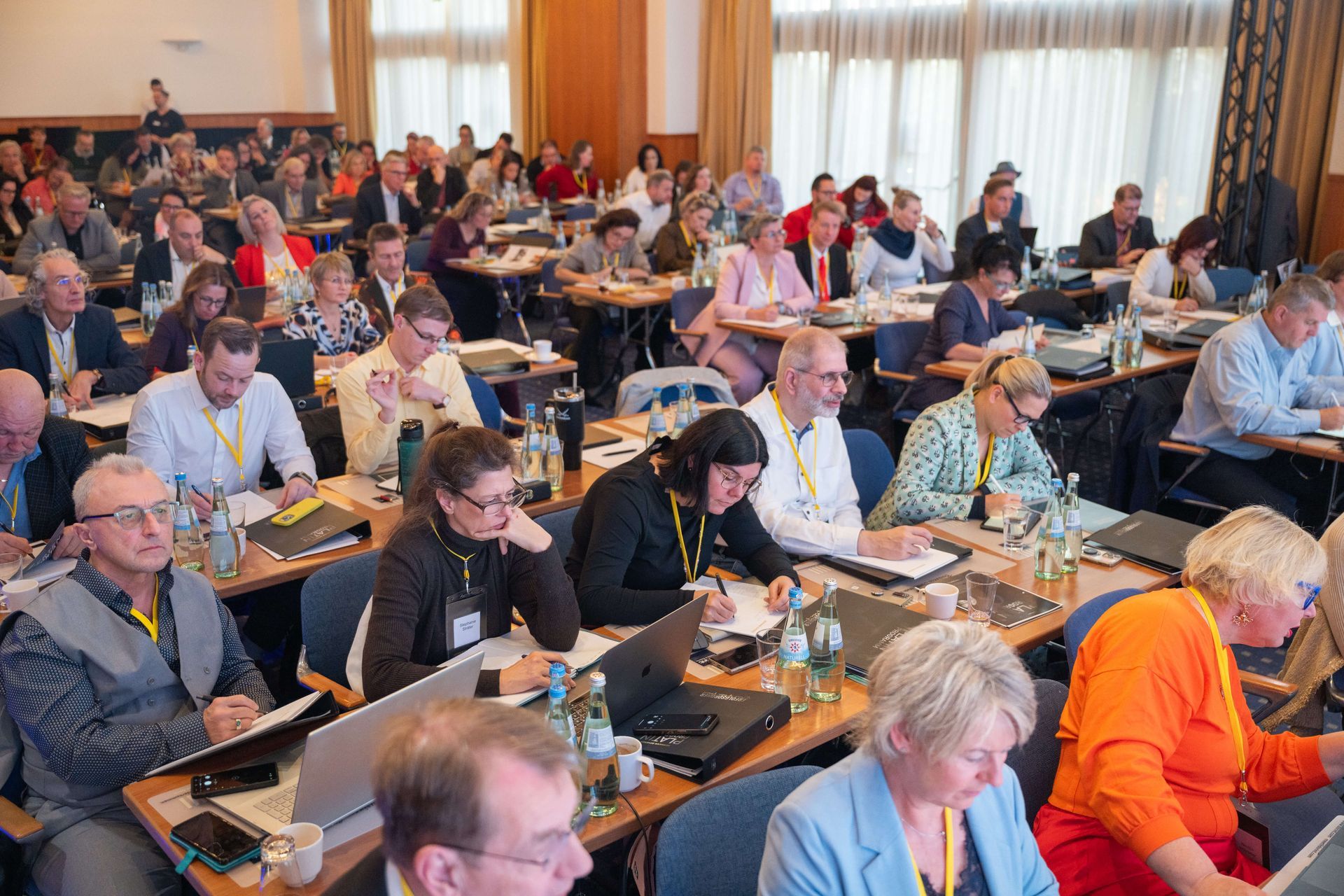 Großer Konferenzraum mit vielen Personen an Tischen, einige machen sich Notizen und benutzen Laptops.