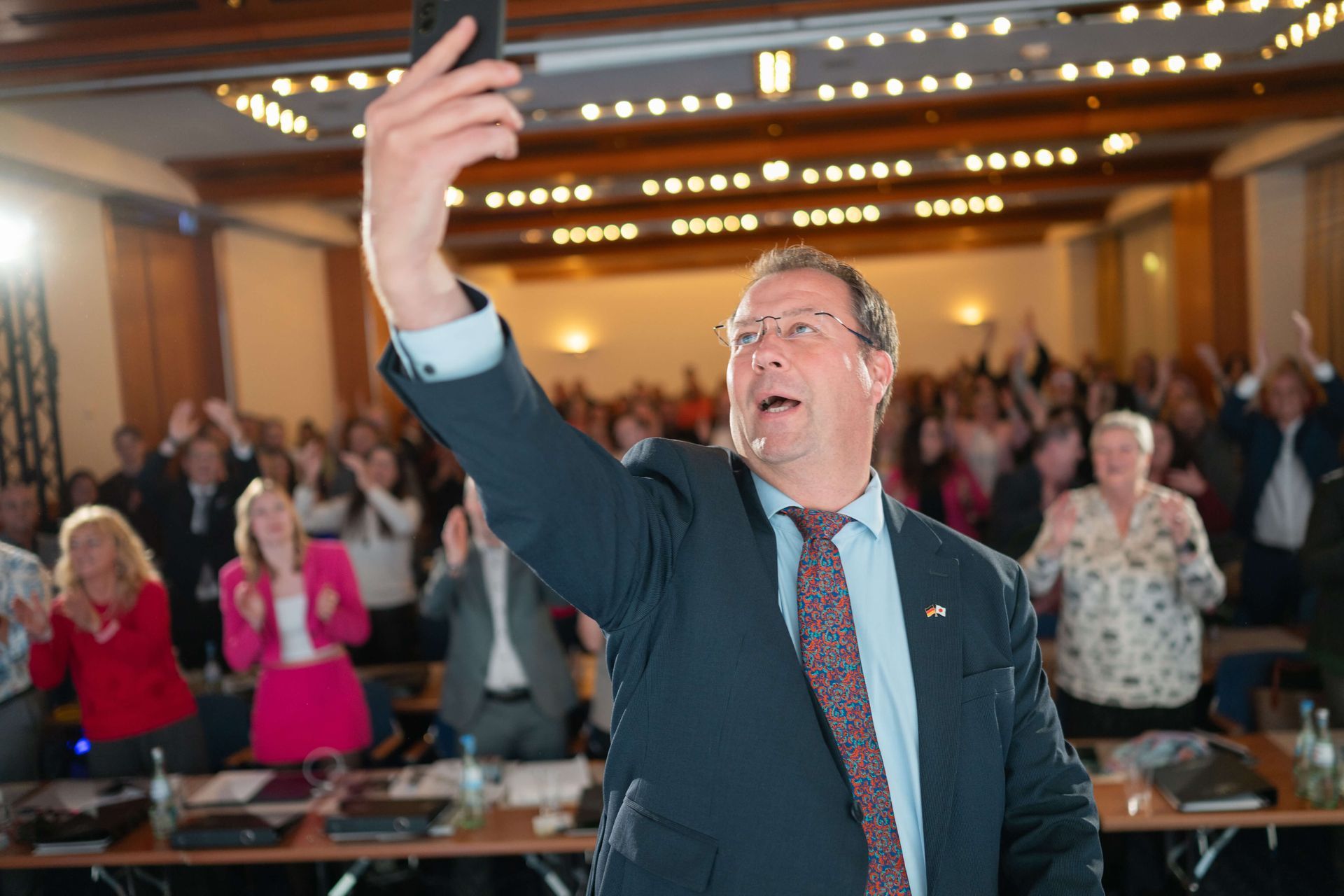 Birger Nispel im Anzug macht ein Selfie mit seinem Handy, im Hintergrund jubelt eine Menschenmenge. Konferenzraum.