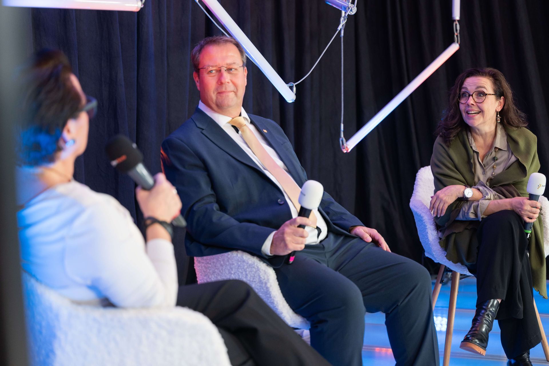 Drei Personen sitzen, jeder hält ein Mikrofon. Birger Nispel im Anzug, zwei Frauen, alle in einer bühnenähnlichen Umgebung mit Beleuchtung.