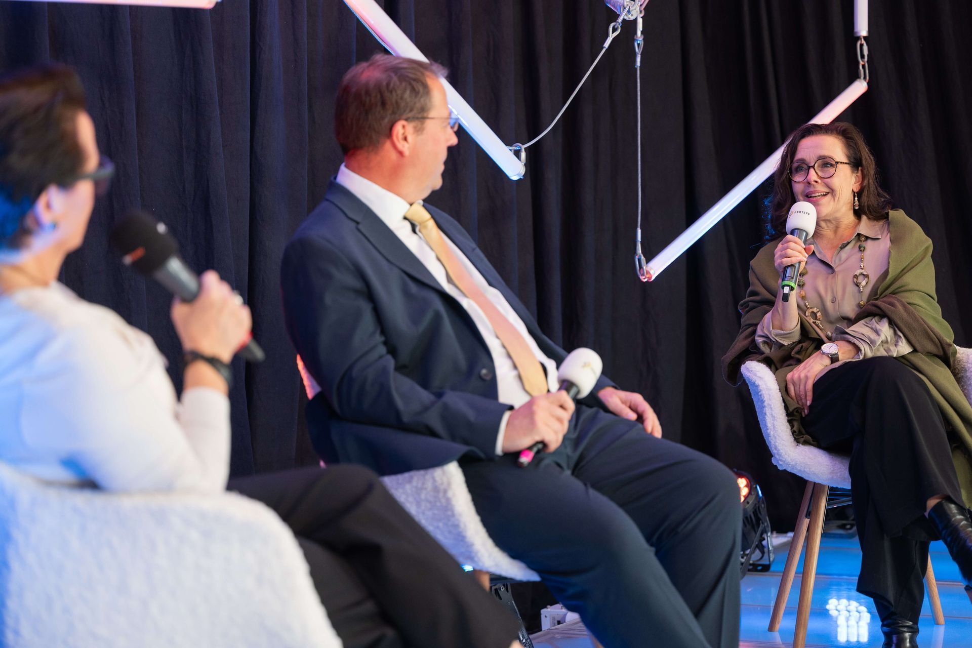 Drei Personen sitzen mit Mikrofonen in der Hand auf der Bühne. Birger Nispel hört zu und eEine Frau spricht ins Mikrofon, zwei andere hören zu.