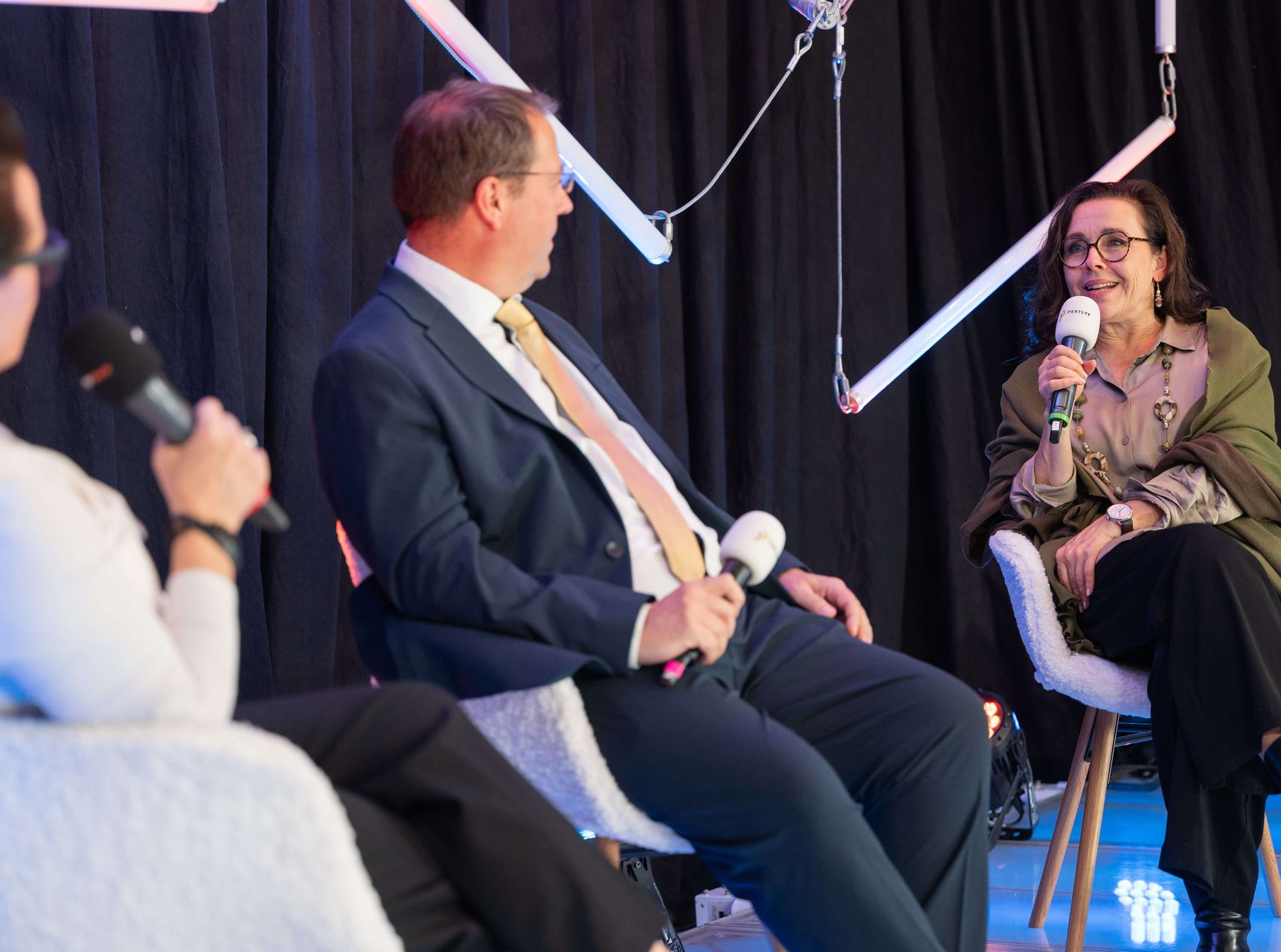 Drei Personen sitzen, halten Mikrofone und sprechen. Birger Nispel im Anzug unterhält sich mit einer Frau. Im Hintergrund ist eine Bühne zu sehen.