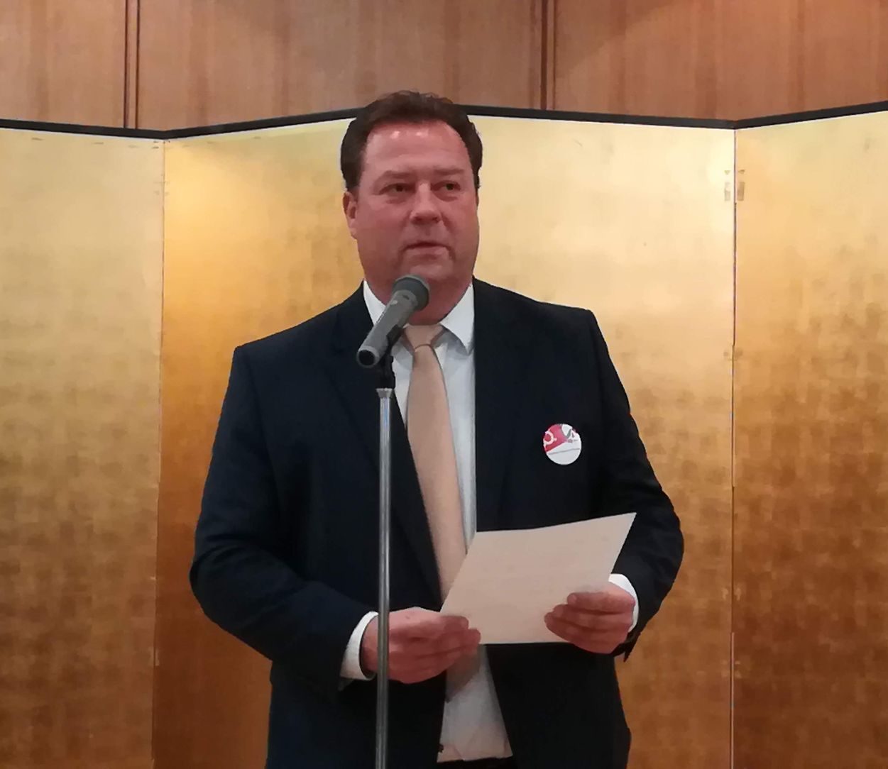 Ein Mann in Anzug und Krawatte steht vor einem Mikrofon und hält ein Stück Papier