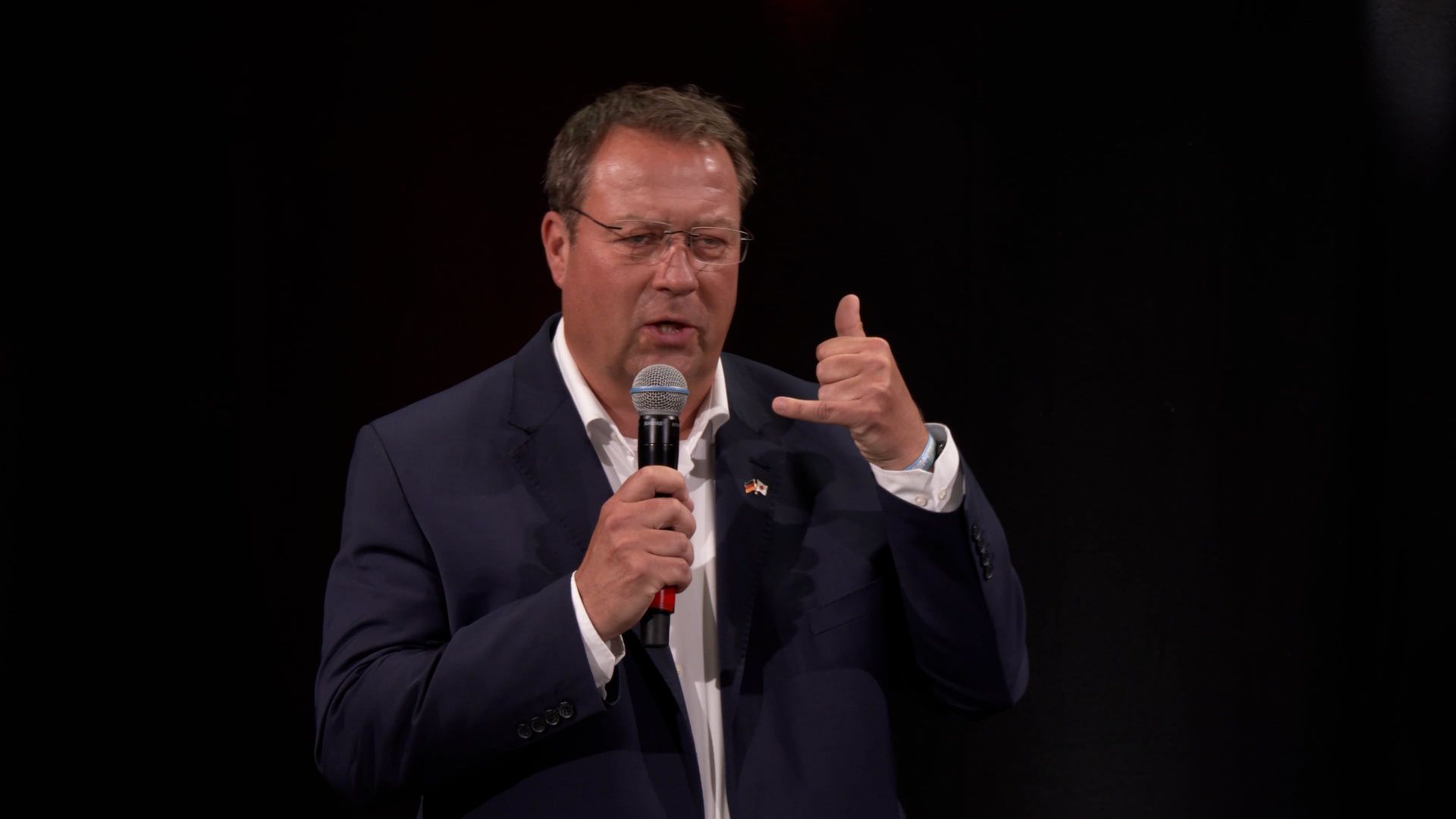 Ein Mann im Anzug hält ein Mikrofon und zeigt mit dem Daumen nach oben.