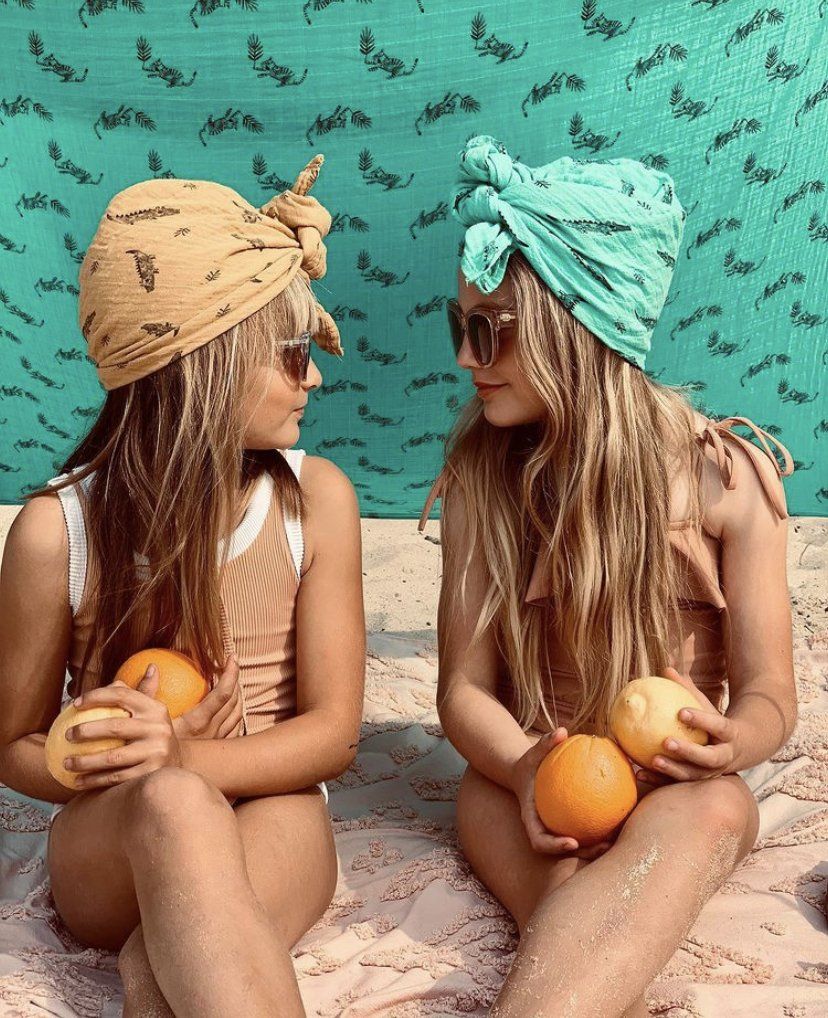 Deux fillettes assises sur la plage avec lunettes de soleil, foulards sur la tête et oranges dans les mains