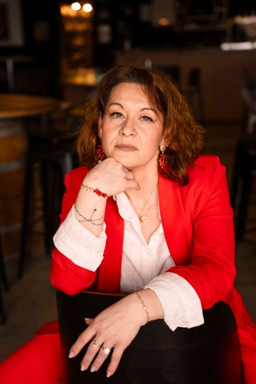Femme en veste rouge, le menton appuyé sur la main. Dans un bar, elle regarde la caméra.