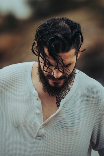 Homme aux cheveux noirs mouillés et à la barbe, portant une chemise blanche en tricot gaufré, regardant vers le bas.