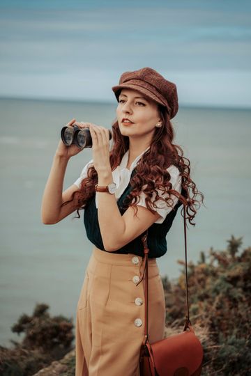 Une femme aux cheveux roux regarde la mer à travers des jumelles, portant une casquette, un gilet et une jupe.