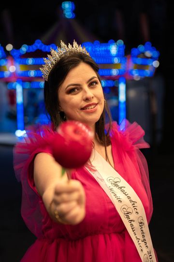 Une femme en robe rose et couronne tient une pomme d'amour, l'écharpe porte l'inscription « Alaharrhice ».