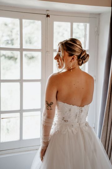 La mariée, vêtue d'une robe bustier, se tient près d'une fenêtre, le regard ailleurs.