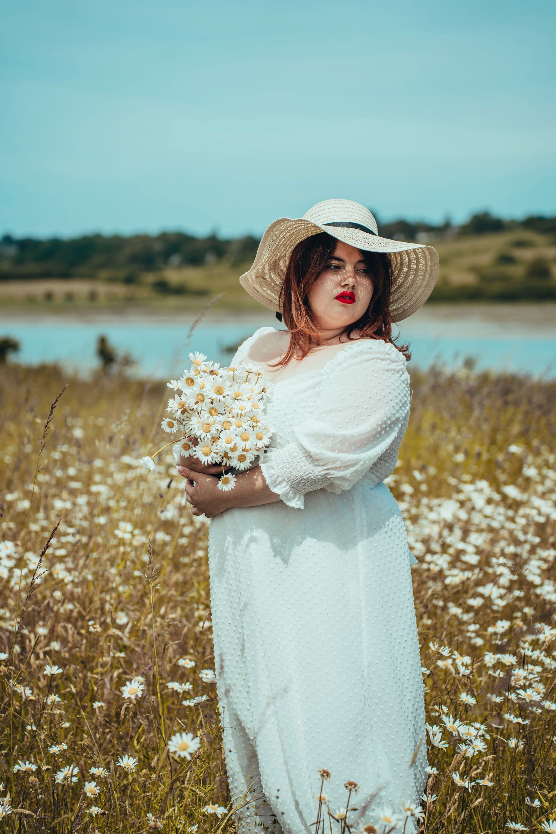 Femme en robe blanche et chapeau de paille tenant des fleurs dans un champ de marguerites, près de l'eau.