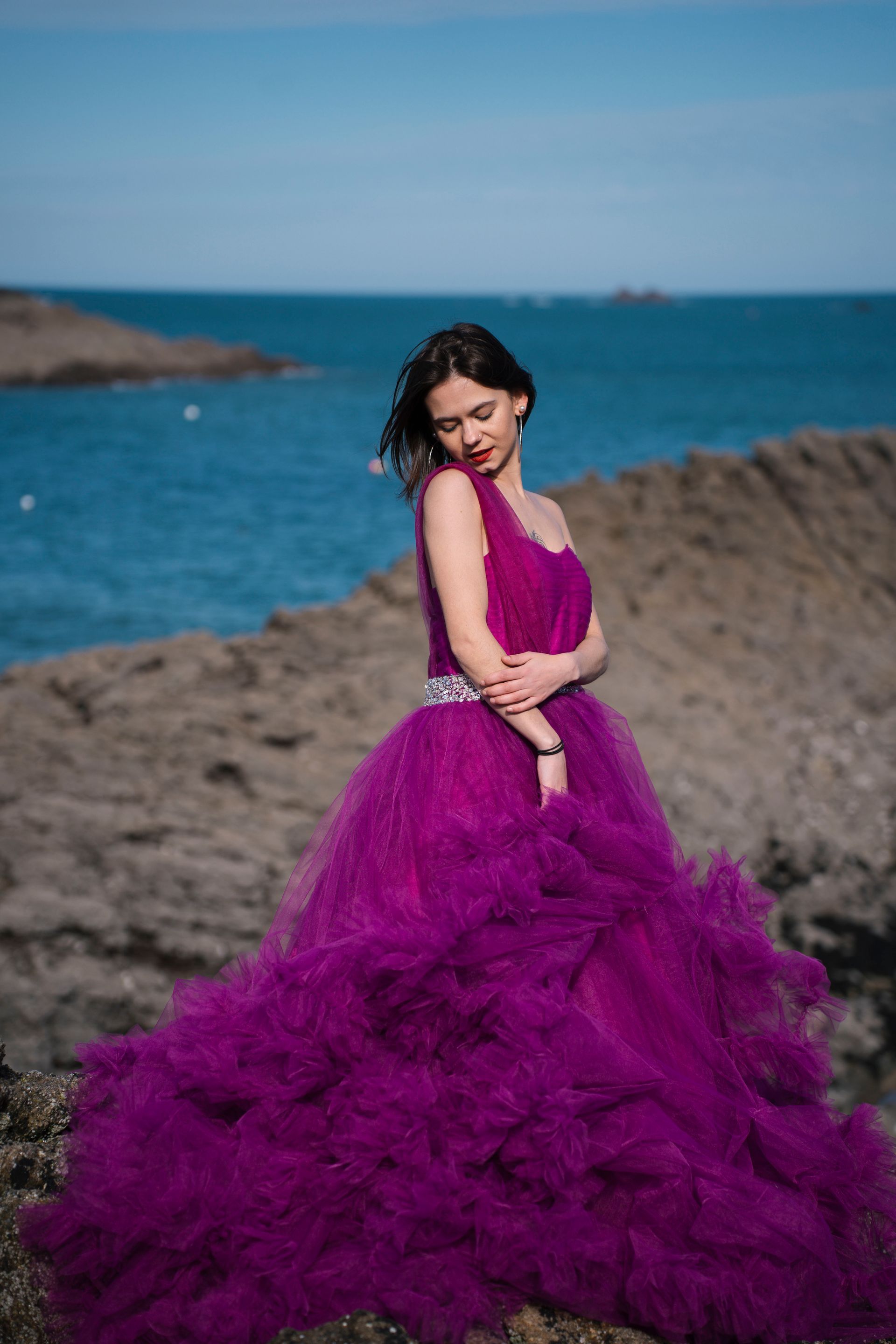 Une femme vêtue d'une robe magenta à une épaule pose sur des rochers au bord de l'océan.
