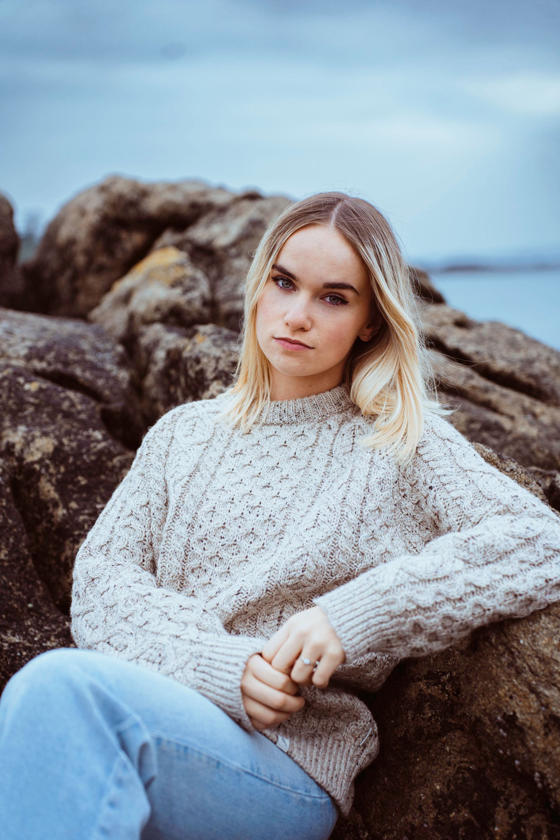Une femme blonde vêtue d'un pull torsadé et d'un jean est assise sur des rochers au bord de l'eau, regardant la caméra.