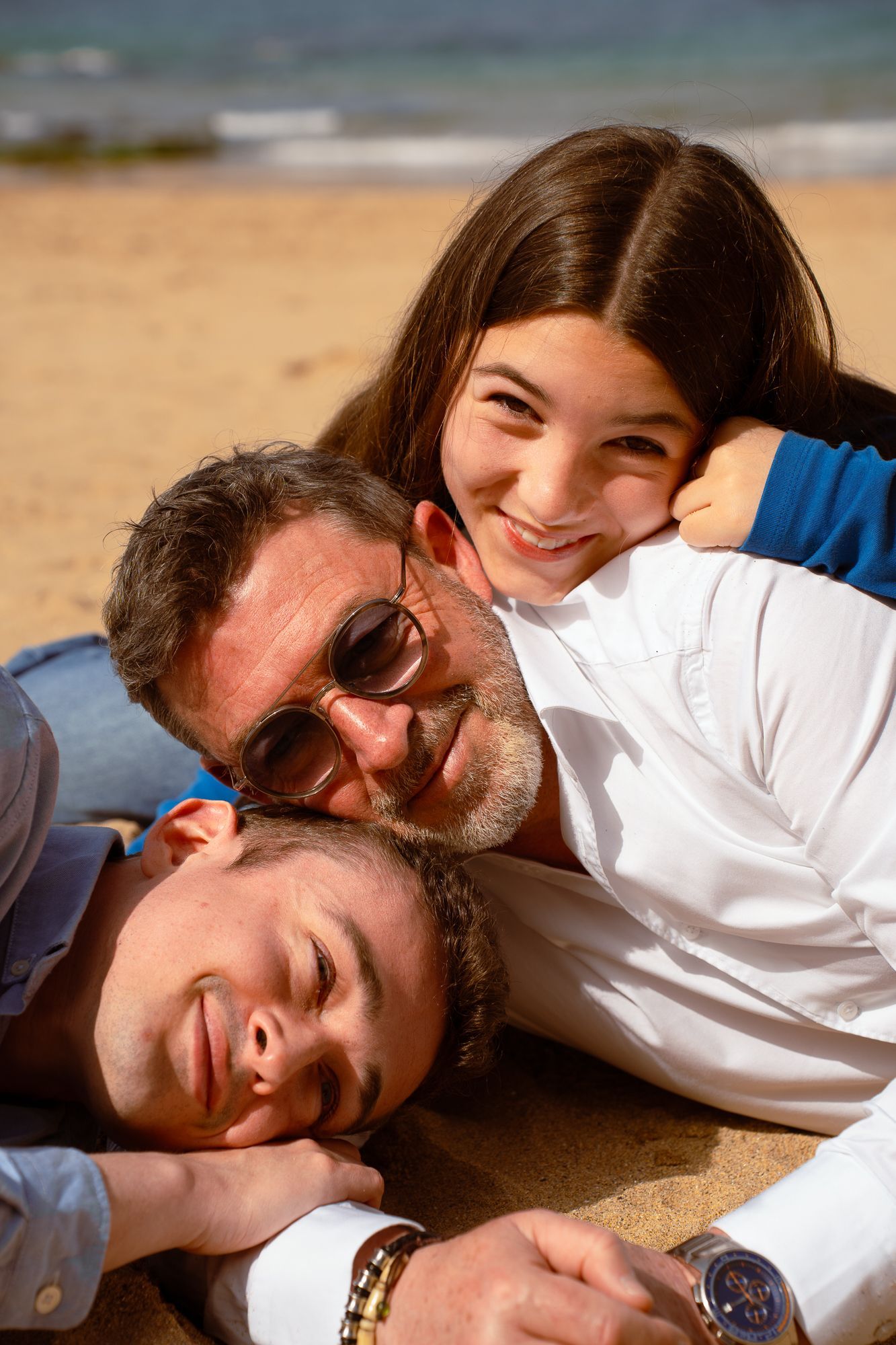 Une famille de trois personnes sur une plage, souriantes et allongées sur le sable. La fille en haut.