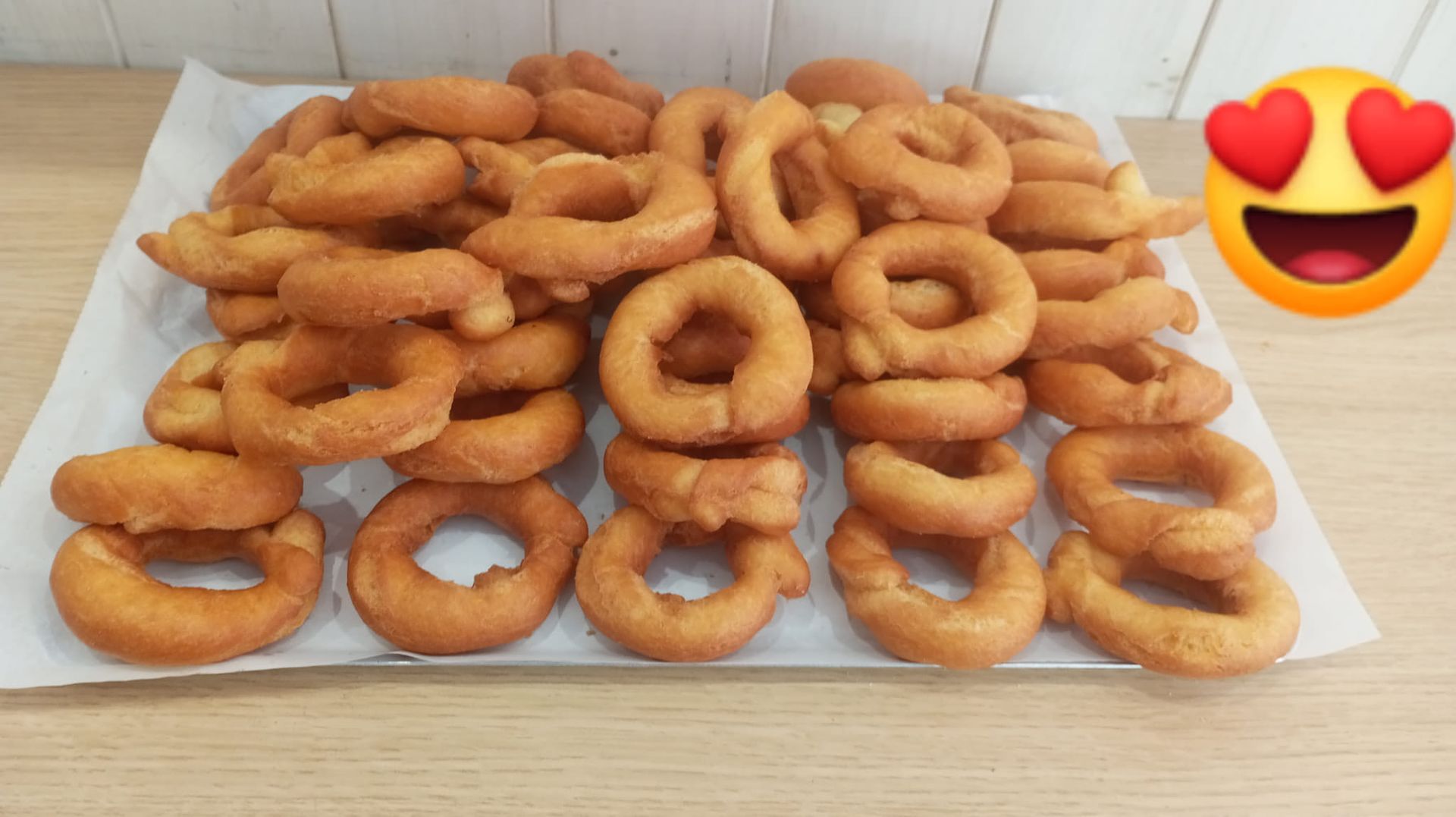 Bandeja de rosquillas doradas en forma de anillo sobre papel pergamino, con un emoji de ojos de corazón en la esquina.
