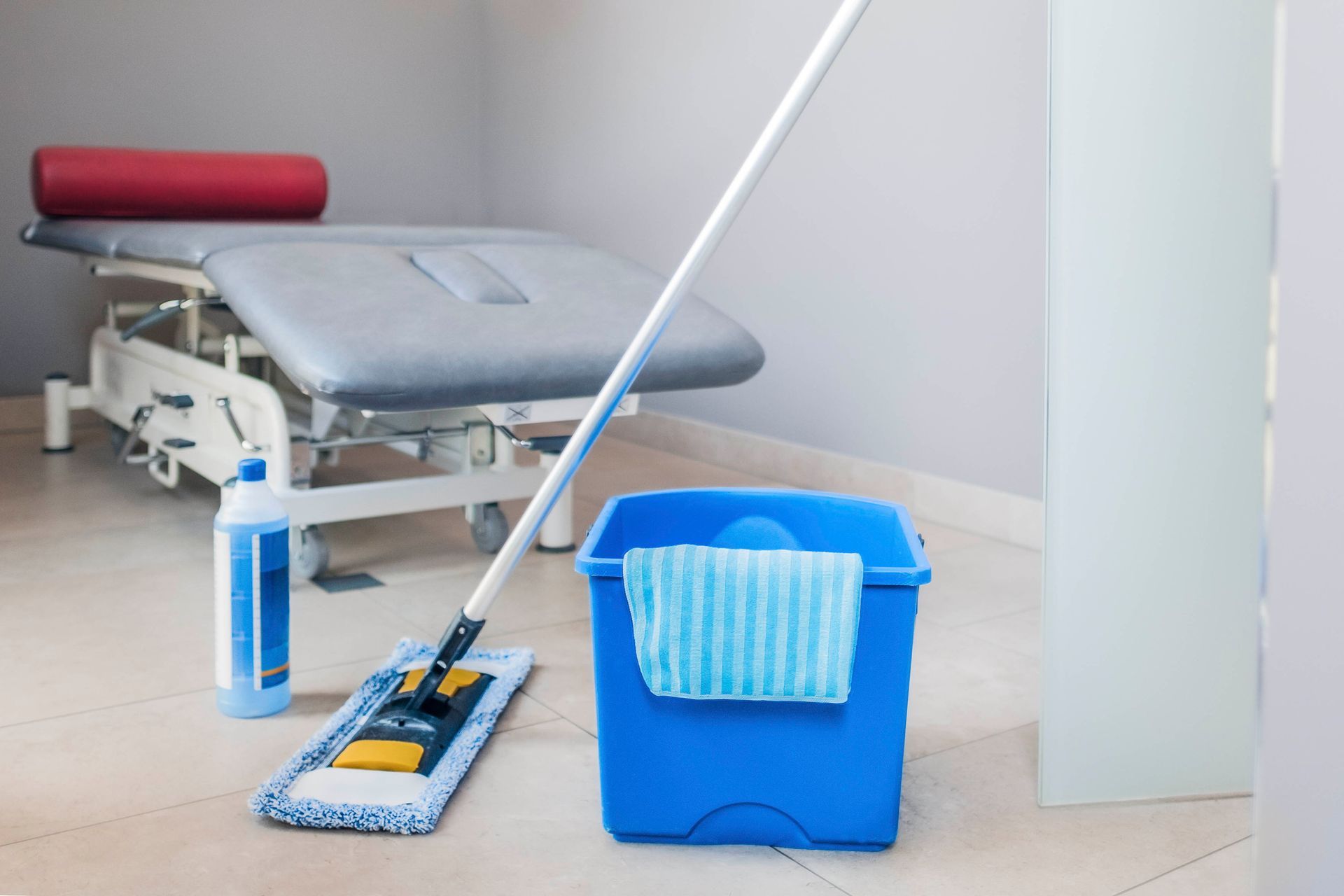 Blauer Mopp und Eimer auf Fliesenboden in einer Klinik, in der Nähe eines Untersuchungstisches.
