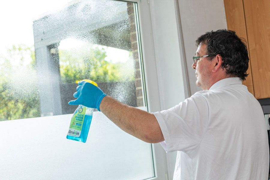 Mann sprüht in einem hellen Raum mit einem blauen Tuch Reinigungsmittel auf eine Fensterscheibe.