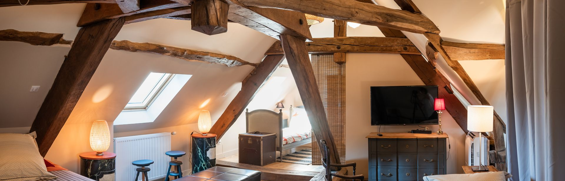 Chambre mansardée avec poutres apparentes, plusieurs lampes, un lit, une télévision et un puits de lumière.