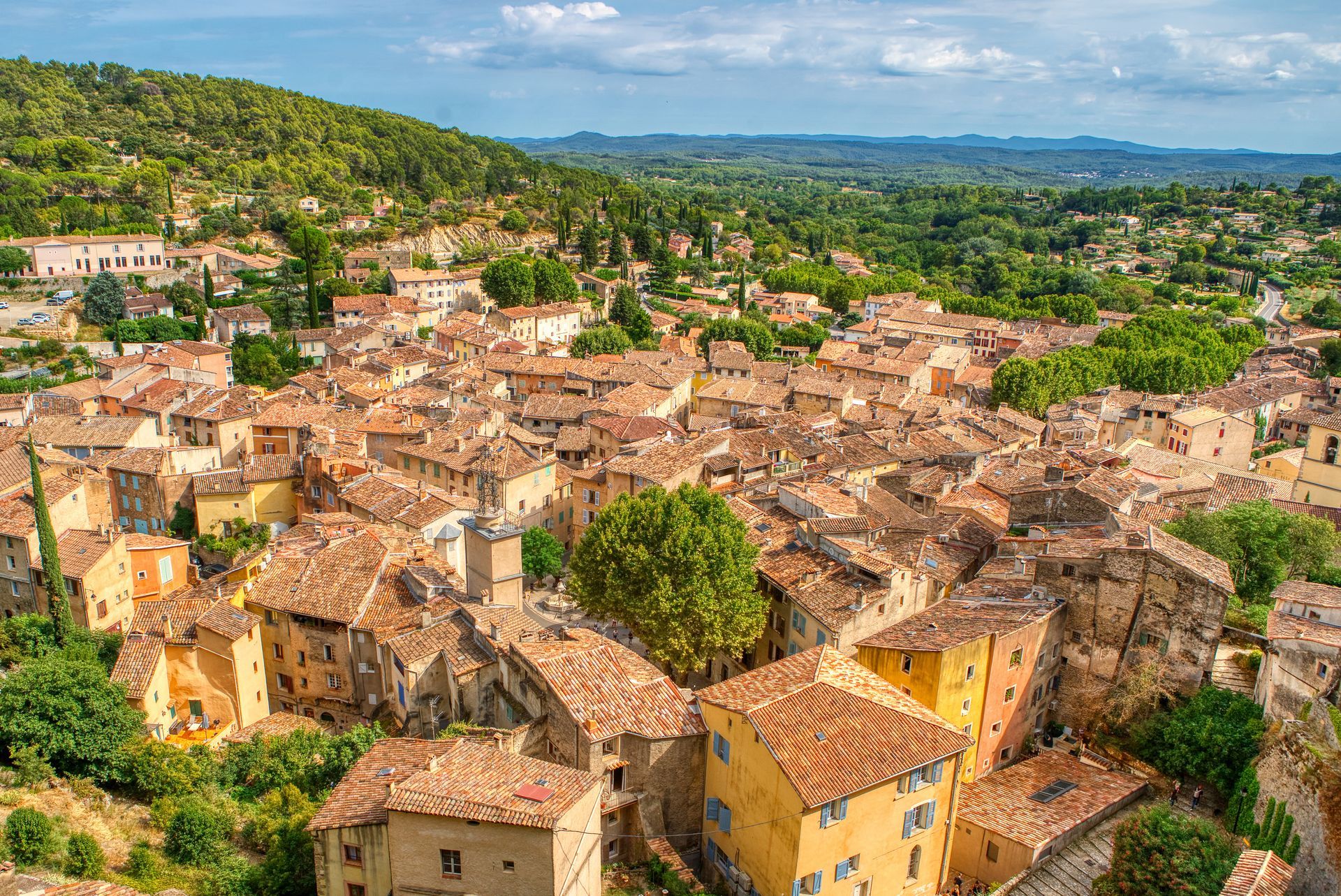 Vue en plongée d'un village provençal aux toits de tuiles en terre cuite, à flanc de colline verdoyante.