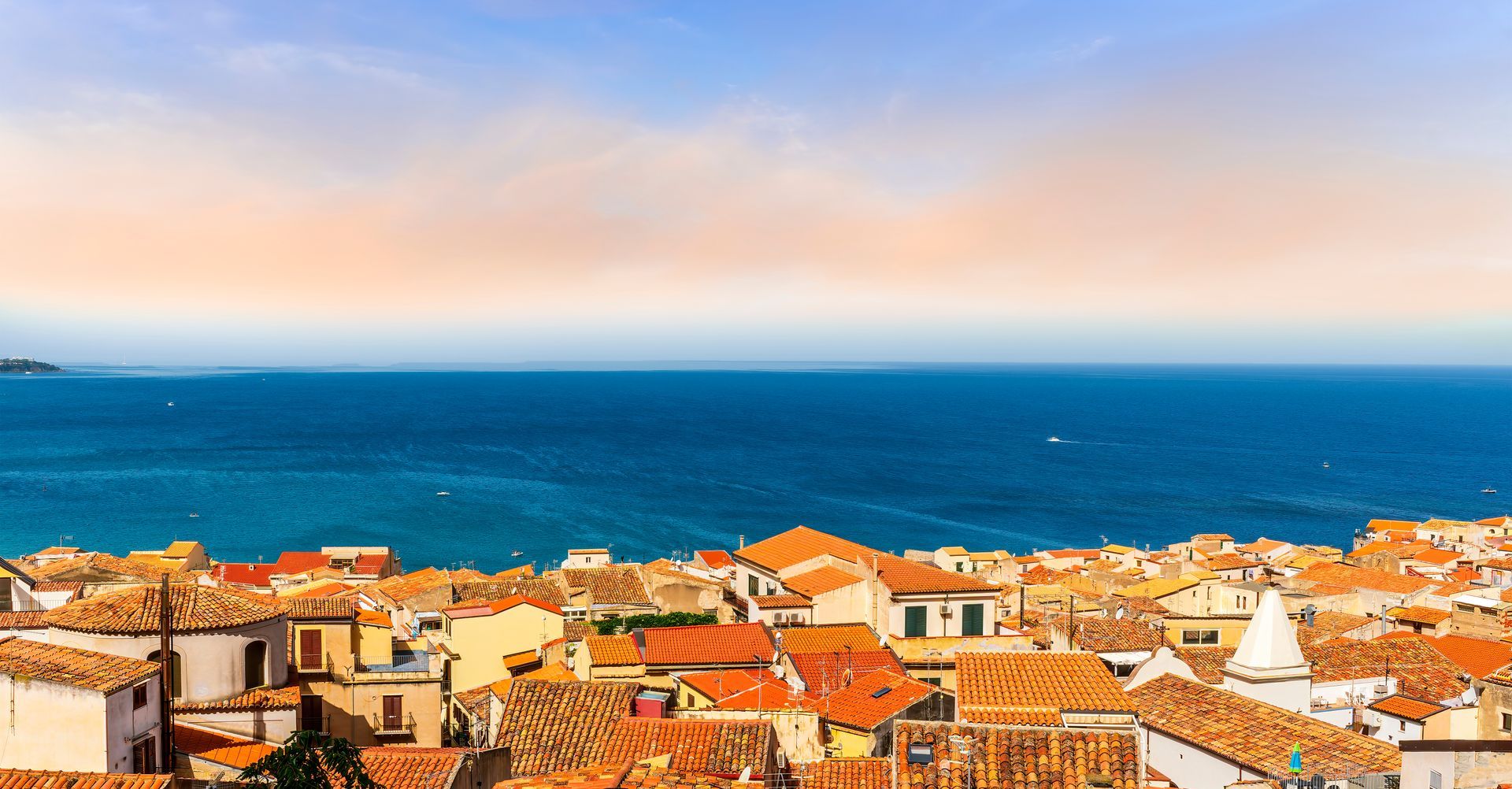 Toits de tuiles rouges d'une ville côtière surplombant une vaste mer d'un bleu profond sous un ciel pâle et brumeux.