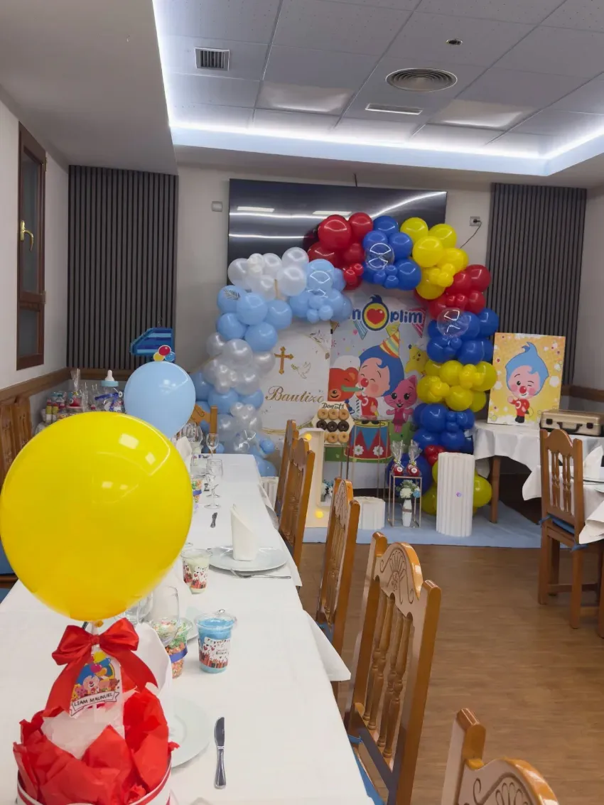 Salón de fiestas decorado con arcos de globos, mesas largas y coloridas exhibiciones de regalos.