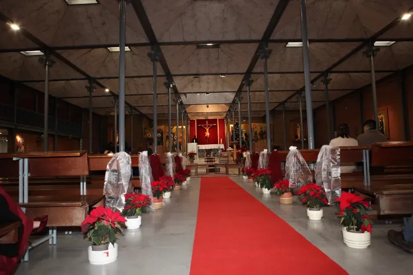 Pasillo con alfombra roja en una iglesia o salón, flanqueado por sillas blancas y arreglos florales rojos.