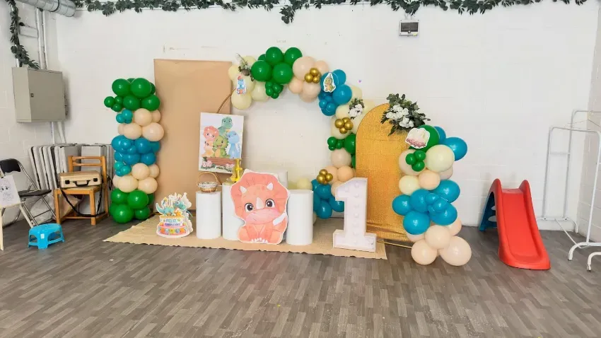 Fondo para fiestas decorado con globos, arcos verdes, dorados y blancos, recortes de dibujos animados y un tobogán rojo a la derecha.