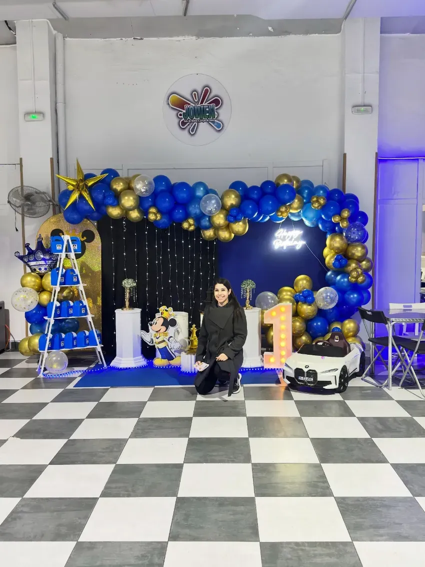 Persona sentada frente a decoraciones de globos azules y dorados sobre un suelo a cuadros, con una torre iluminada cerca.