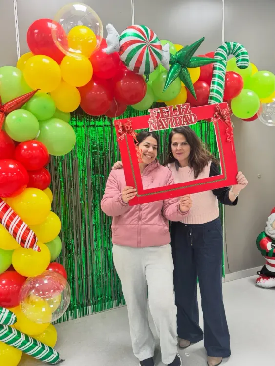 Dos mujeres posando con un marco de fotos rojo frente a un colorido fondo de fotomatón con globos.