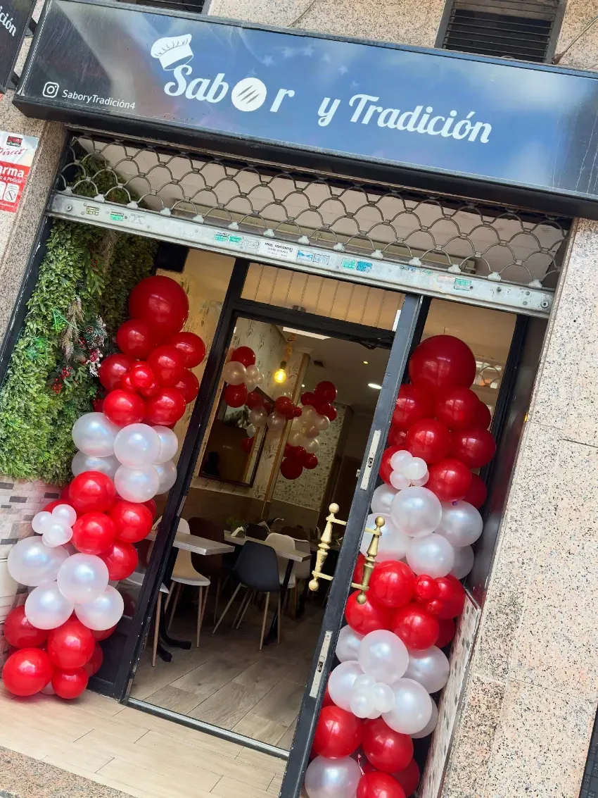 Fachada decorada con columnas de globos rojos y blancos alrededor de una entrada de cristal bajo un letrero azul.
