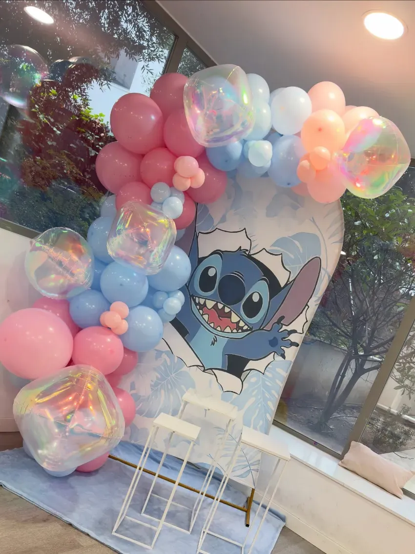 Arco de globos en tonos pastel que enmarca un fondo de Stitch y sillas blancas en un espacio para eventos decorado.