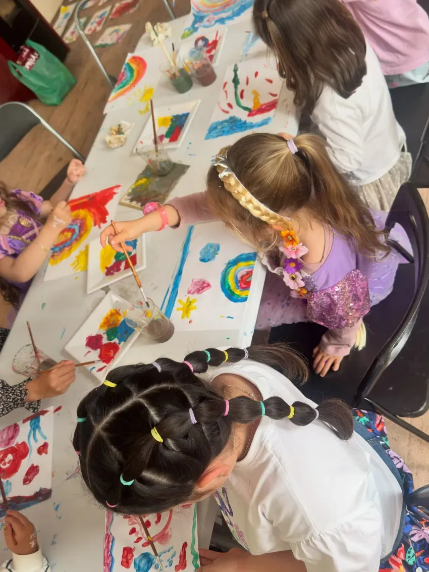 Niños pintando coloridas obras de arte en papel en una mesa del aula con pinceles y vasos de pintura.