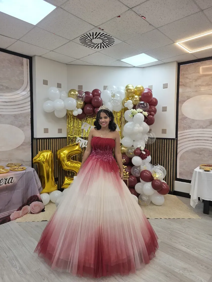 Una persona con un vestido de gala rojo y blanco se encuentra de pie frente a un fondo de globos dorados y blancos en una celebración.