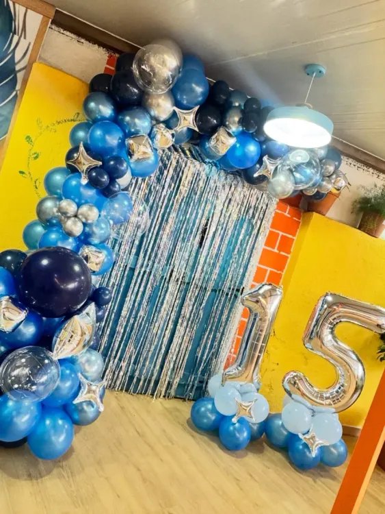 Arco de globos azules y plateados que enmarca una cortina brillante, con un gran globo plateado número 15 a la derecha.