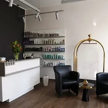 Ein Salonempfangsbereich mit weißem Schreibtisch, Produktregalen, zwei schwarzen Ledersesseln und einem goldenen Gepäckwagen.