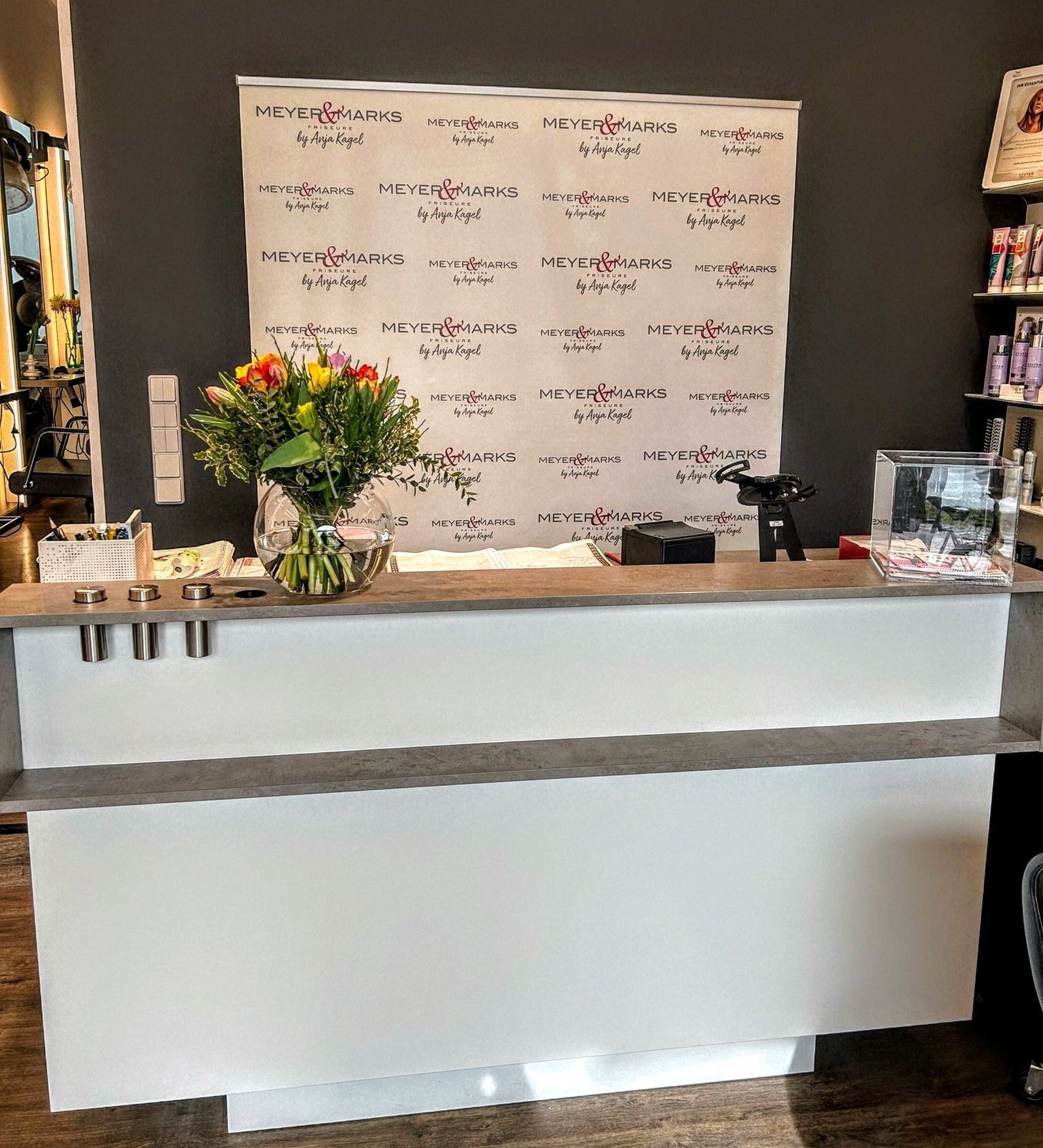 Empfangstresen vom Friseursalon Kagel in Werder mit einem Blumenarrangement und dem Firmenlogo im Hintergrund.