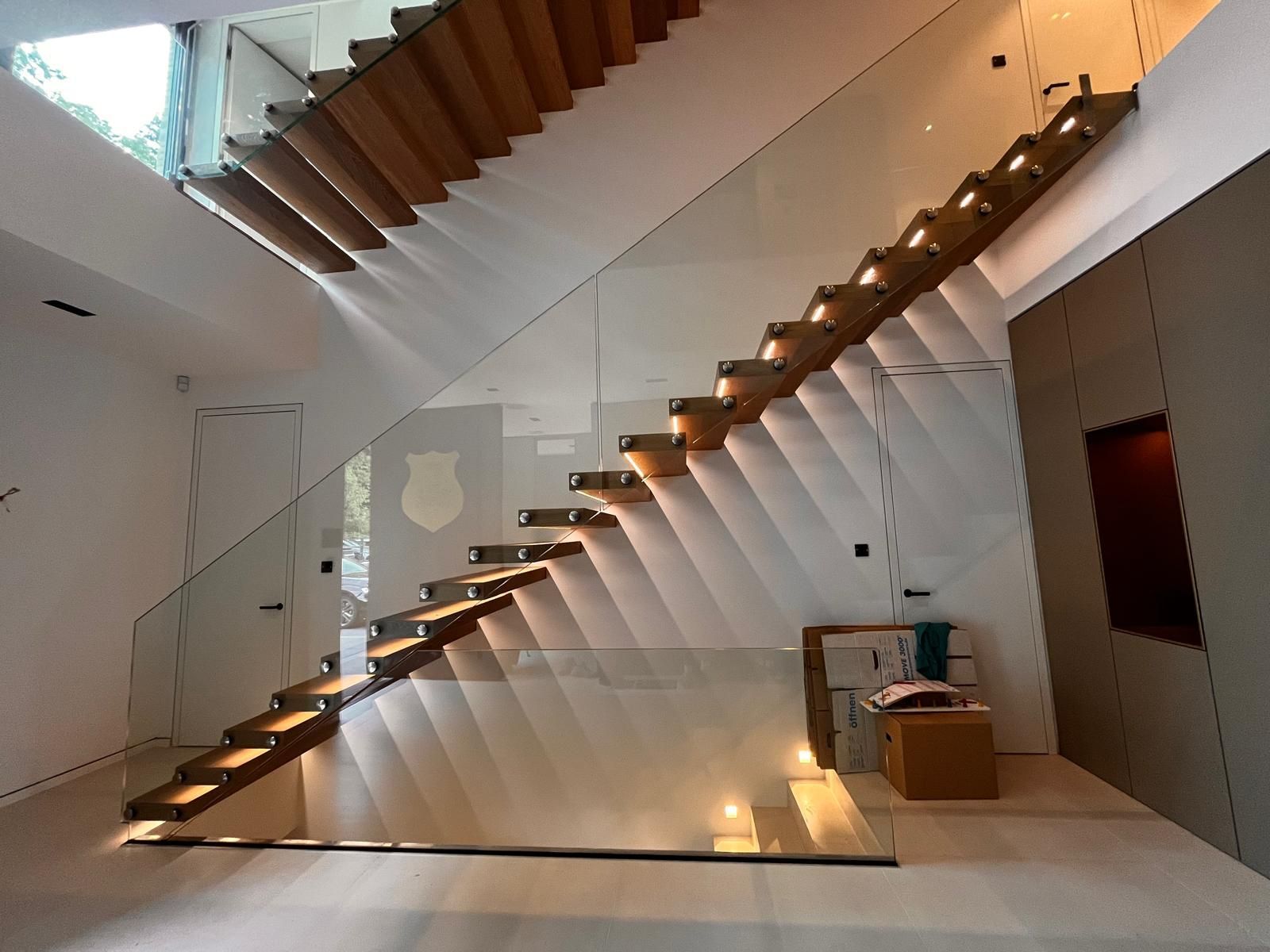 Modernes, weißes Interieur mit langer Holztreppe und Glaswänden, beleuchtet von warmen Strahlern.