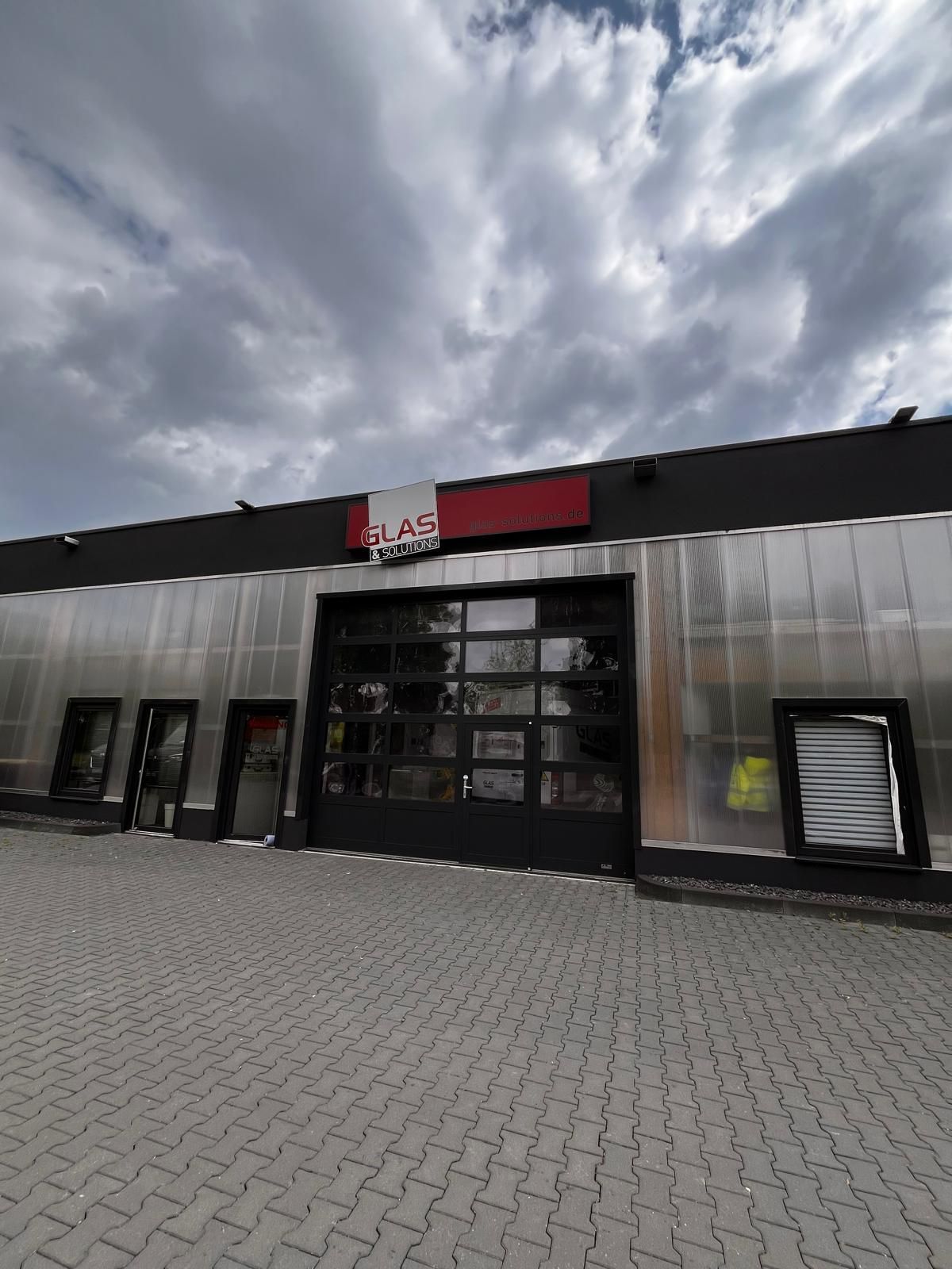 Moderne Gebäudefassade mit Glastüren, rotem Schild und bewölktem Himmel über einem gepflasterten Vorplatz