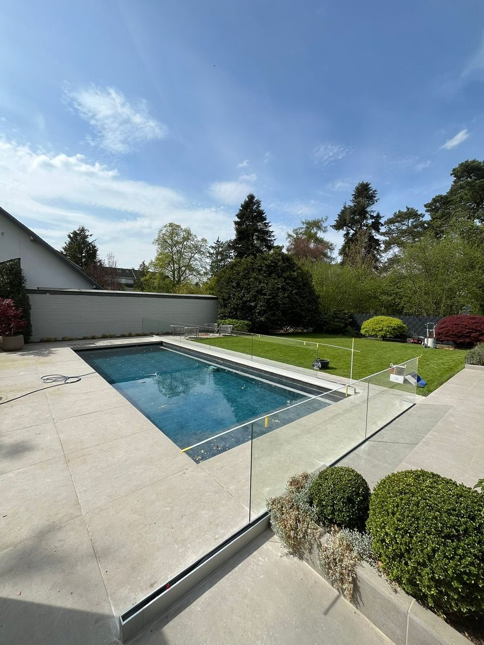 Hinterhofterrasse mit rechteckigem Swimmingpool, grünem Rasen und gepflegten Sträuchern unter blauem Himmel