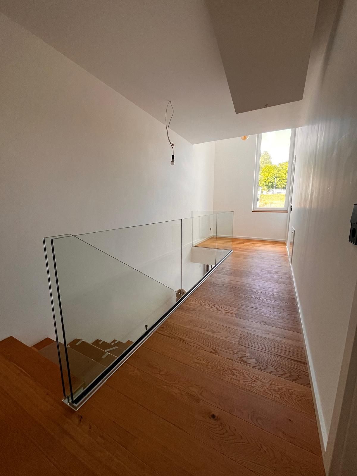 Schmaler Flur mit Holzboden, weißen Wänden, Glasgeländer und einem Fenster am Ende.