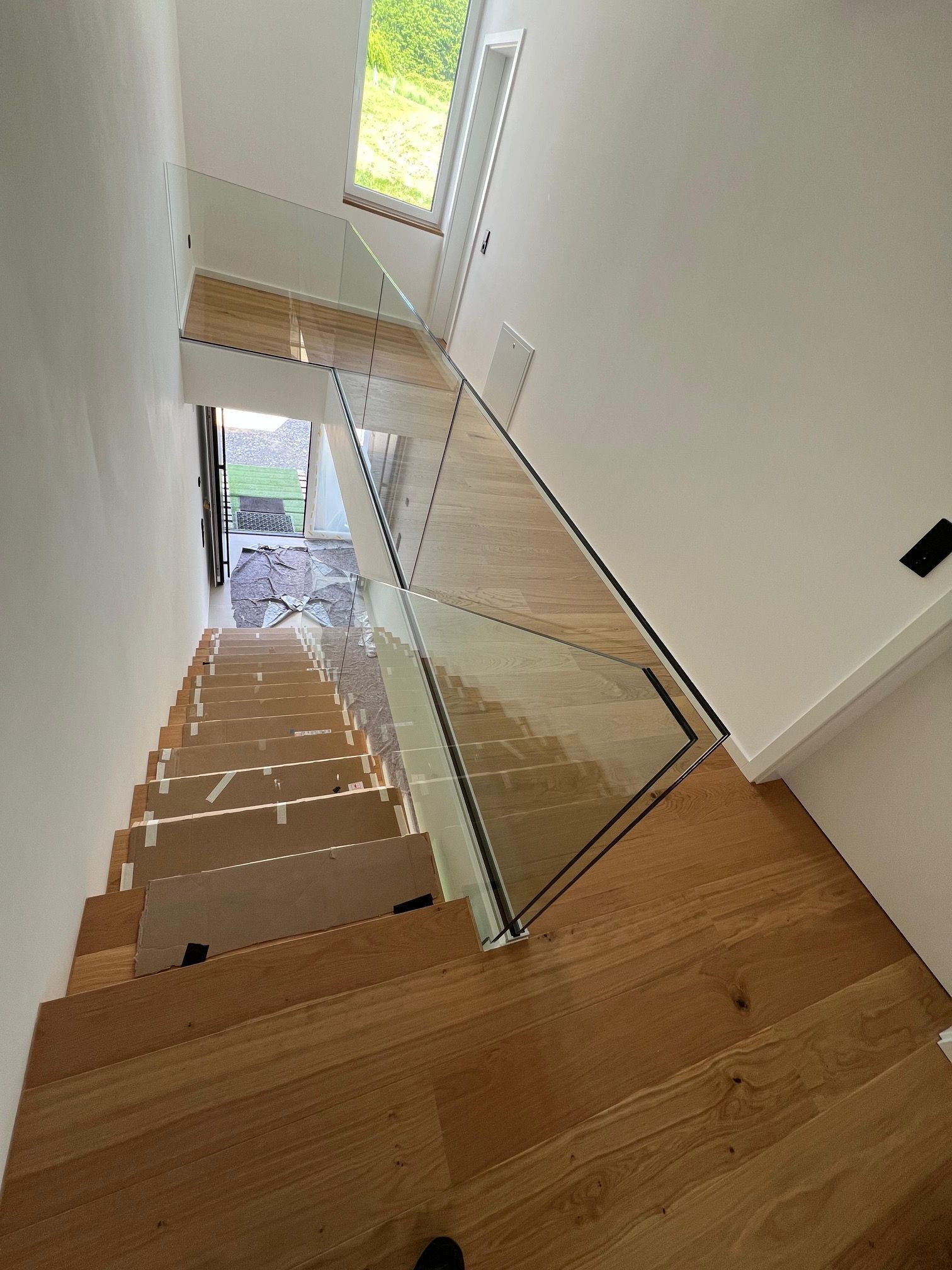 Blick hinunter über eine schmale Holztreppe mit Glasgeländer, hinab zu einem hellen Türrahmen und Eingang.